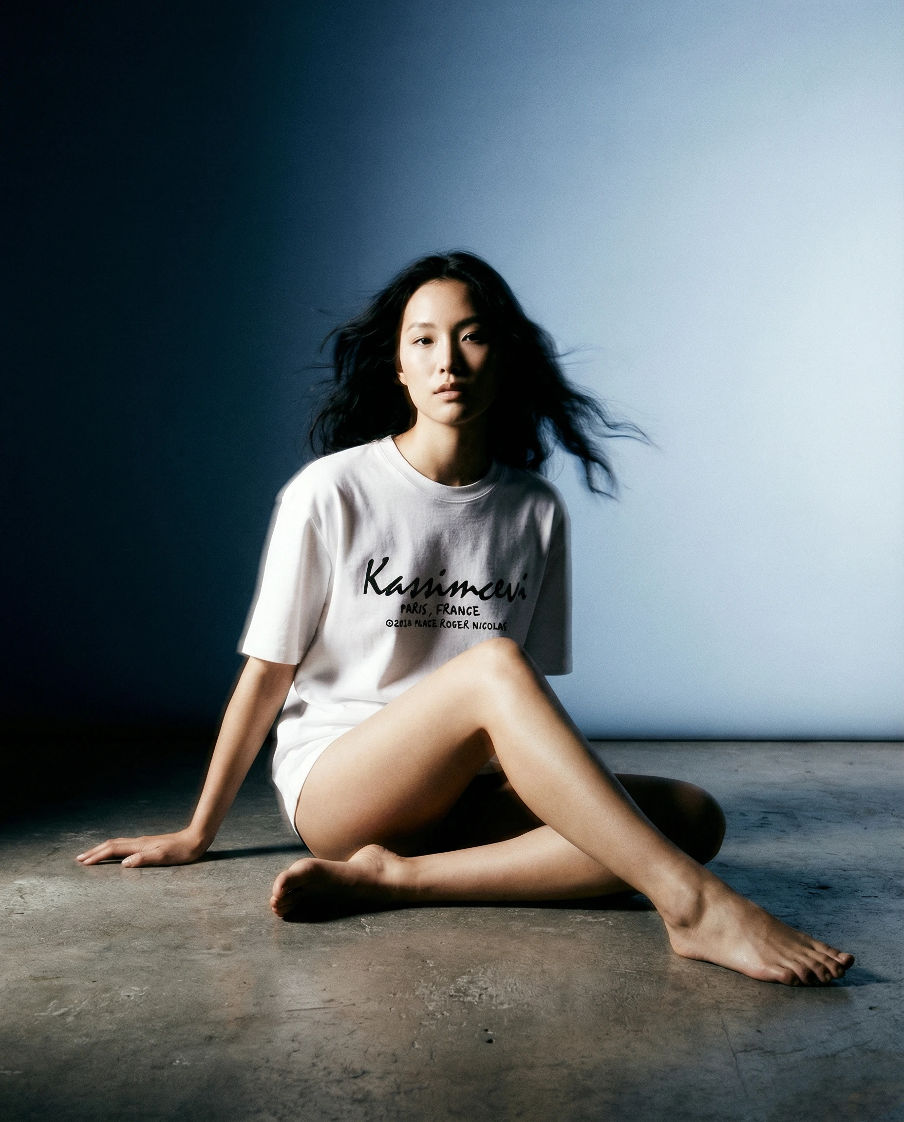 Seated model wearing the Place Roger Nicolas × Kassimcevi white T-shirt against a blue backdrop